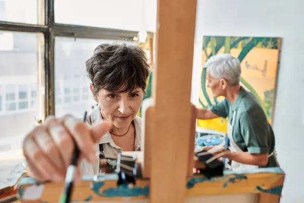 Brunette mature woman near easel and female girlfriend painting on blurred backdrop, artistic hobby — Stock Photo
