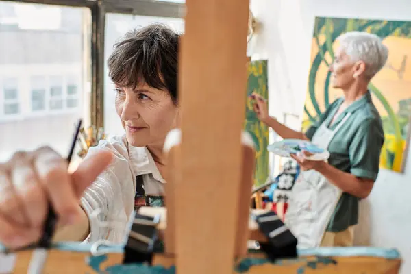 Creative mature woman near easel and female girlfriend painting on blurred backdrop, artistic hobby — Stock Photo