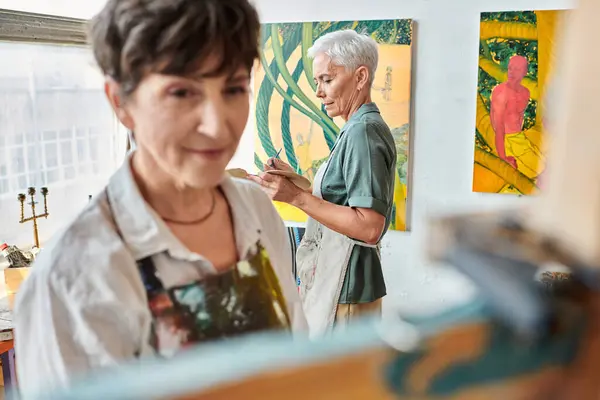 Blurred female artist near easel and mature woman painting in art workshop, creative hobby — Stock Photo