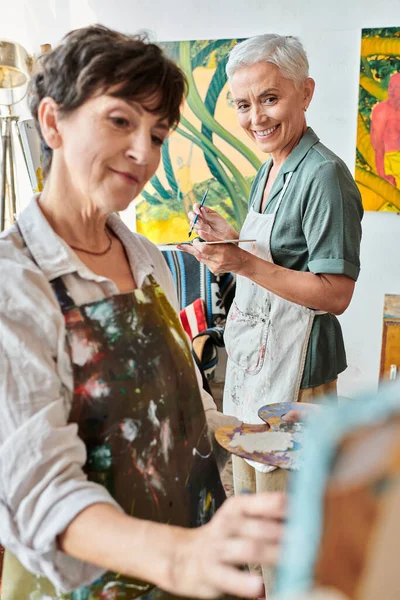 Cheerful mature woman in apron looking at female friend painting on blurred foreground, creativity — Stock Photo