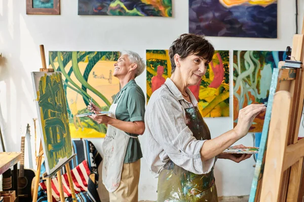 Joyful stylish mature women in aprons pointing on easels in art workshop, friendship and creativity — Stock Photo