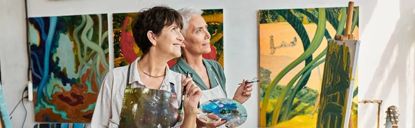 Joyful creative mature women in aprons looking away in modern art workshop, horizontal banner — Stock Photo