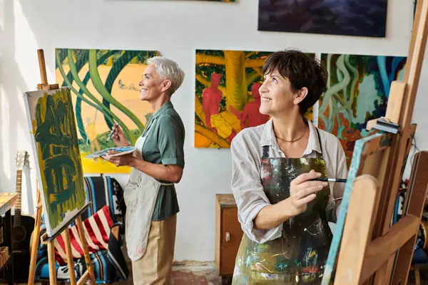 Smiling mature women in aprons painting on easels and looking away in art workshop, creative hobby — Stock Photo