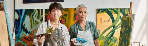 Талановиті жінки-художники в фартухах тримають палітру і дивляться на камеру в художній студії, банер — стокове фото