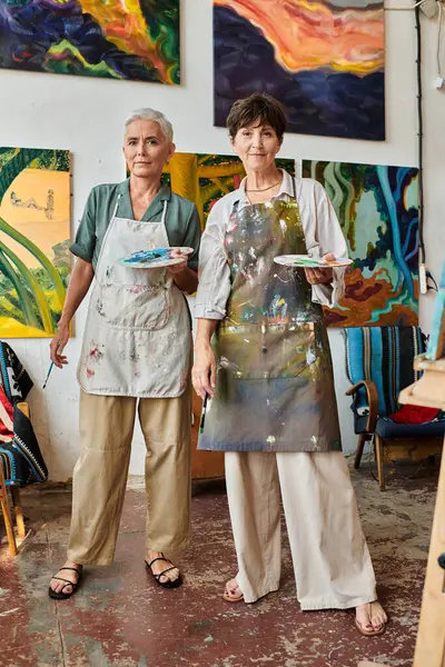 Creative mature female friends holding palettes and looking at camera in art studio, artistic hobby — Stock Photo