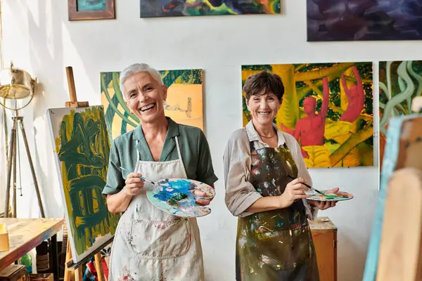 Excitadas amigas maduras sosteniendo paletas y sonriendo a la cámara en el estudio de arte, hobby artístico - foto de stock