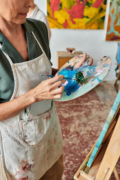 Cropped view of mature female artist mixing on colors on palette with brush in art workshop — Stock Photo