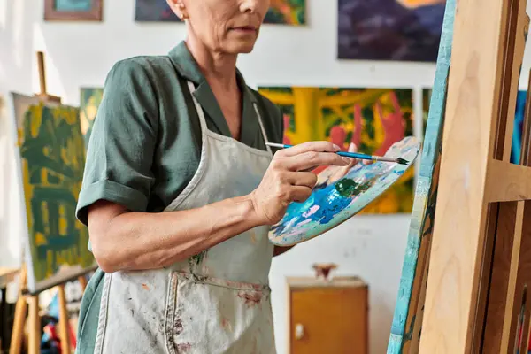 Vue partielle de la femme mature dans le tablier avec palette et peinture au pinceau sur chevalet dans l'atelier d'art — Photo de stock