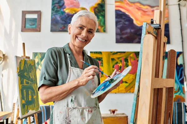Cheerful mature woman with paintbrush and palette looking at camera near easel in art studio — Stock Photo