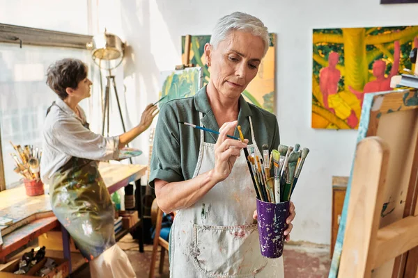 Зріла жінка в фартусі вибирає пензлик біля жіночого друга живопису в художній студії, творче хобі — стокове фото