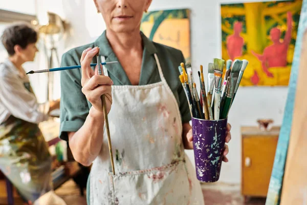 Cropped view of mature woman with paintbrushes near female friend painting in art workshop — Stock Photo