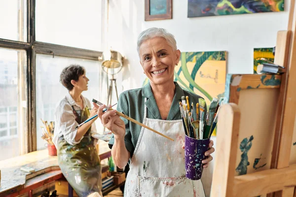 Joyful mature woman with set of paintbrushes near female friend in art workshop, creative hobby — Stock Photo