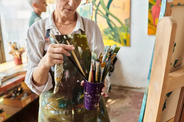 Cropped view of mature woman choosing paintbrushes near female friend painting in art workshop — Stock Photo