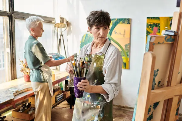 Talented mature woman with paintbrushes looking at easel near female friend in art workshop — Stock Photo
