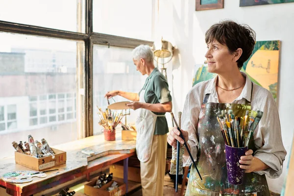 Smiling middle aged woman with set of paintbrushes looking away near female friend in art studio — Stock Photo