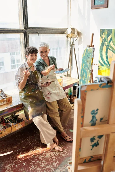 Smiling mature female artists pointing at easel with paintbrush craft workshop, creative hobby — Stock Photo