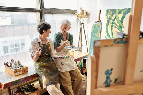 Взволнованные зрелые художницы с палитрами и кистями разговаривают возле мольберта в мастерской — стоковое фото