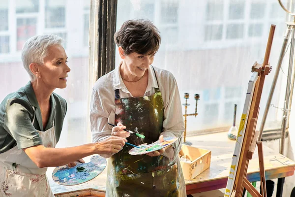 Mature female artists mixing colors on palettes near easel in craft workshop, painting master class — Stock Photo
