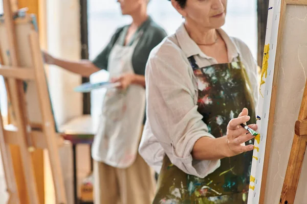 Cropped view of mature woman artist in apron near easel in craft workshop, painting master class — Stock Photo
