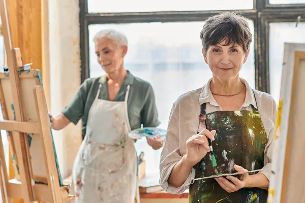Cheerful mature woman holding color palette and smiling at camera during master class in art studio — Stock Photo