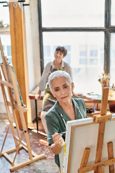 Inspired mature woman in apron painting on easel at master class in art workshop, creative hobby — Stock Photo