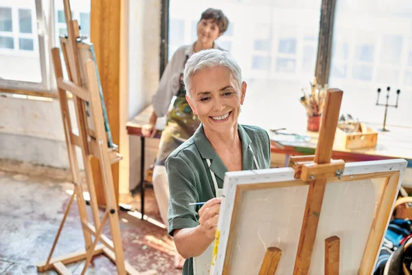 Cheerful mature woman painting on easel near skilled female artist in workshop, master class — Stock Photo