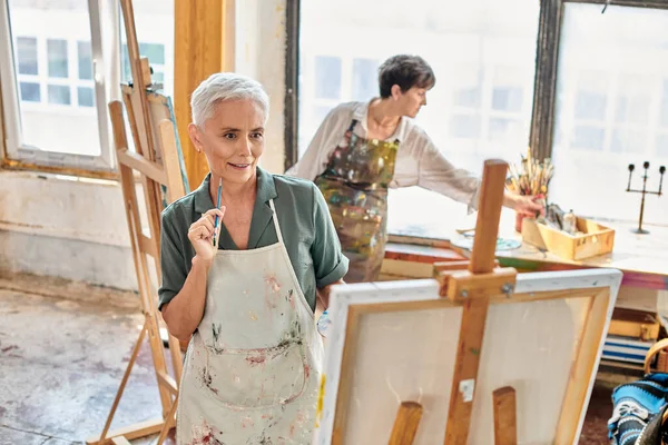 Femme mûre réfléchie regardant chevalet près artiste féminine qualifiée en atelier, classe de maître — Photo de stock