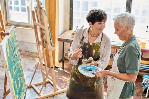 Artiste féminine montrant comment mélanger des peintures sur palette de couleurs à la femme mûre dans un studio d'art, classe de maître — Photo de stock