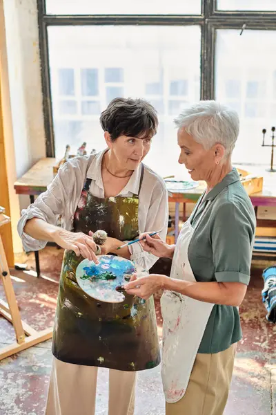 Female artist mixing paints on color palette near mature woman in art studio, painting master class — Stock Photo