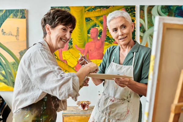 Sorridente donne mature con tavolozza di colori guardando cavalletto durante la pittura master class in studio d'arte — Foto stock