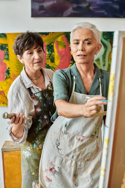 Mature female artist pointing at easel near woman during master class in modern craft workshop — Stock Photo