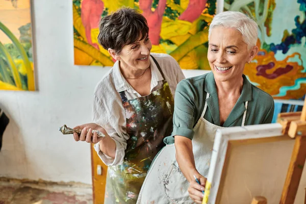Happy middle aged woman painting on easel near skillful female artist during master class in studio — Stock Photo