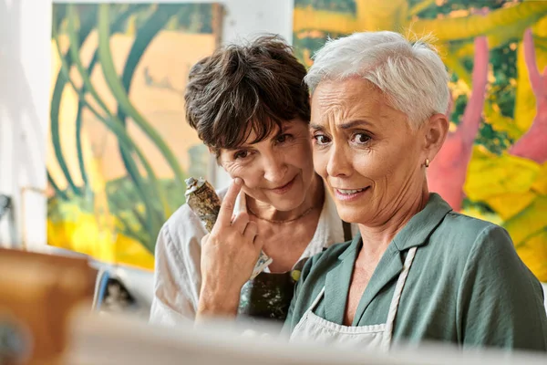 Positive and pensive mature women looking at easel during painting master class in art workshop — Stock Photo