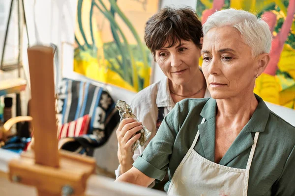 Thoughtful mature women looking at easel during painting master class in modern craft workshop — Stock Photo