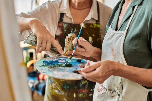 Cropped view of mature women in aprons mixing paints on palette during master class in art workshop — Stock Photo