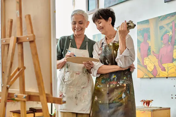 Mulher madura alegre misturando tinta na paleta perto de sorrir artista feminino, master class no estúdio de arte — Fotografia de Stock