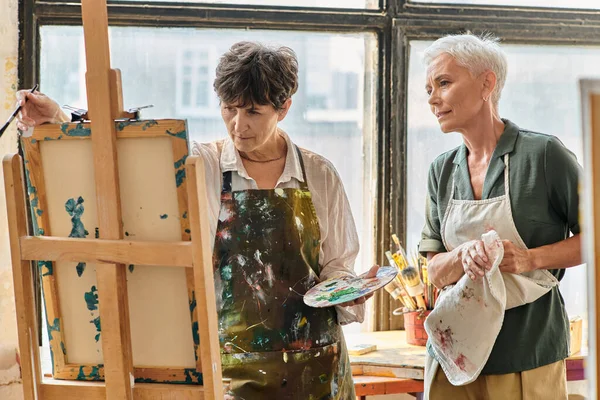Thoughtful mature woman looking at easel near skilled female artist during master class in studio — Stock Photo
