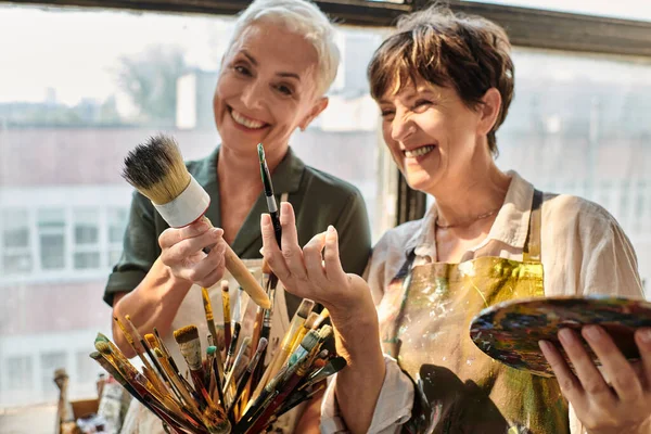 Cheerful mature female women holding different paintbrushes during master class in art studio — Stock Photo
