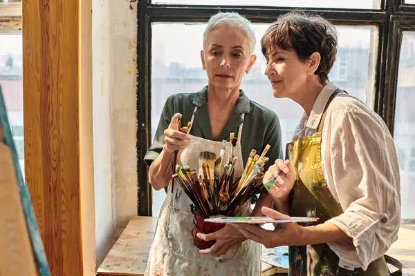 Hábil artista femenina ayudando a las mujeres maduras a elegir el pincel durante la clase magistral en el estudio de arte - foto de stock