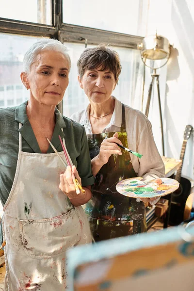 Mujeres de mediana edad artistas con pinceles mirando borrosa caballete durante la clase magistral en el estudio - foto de stock