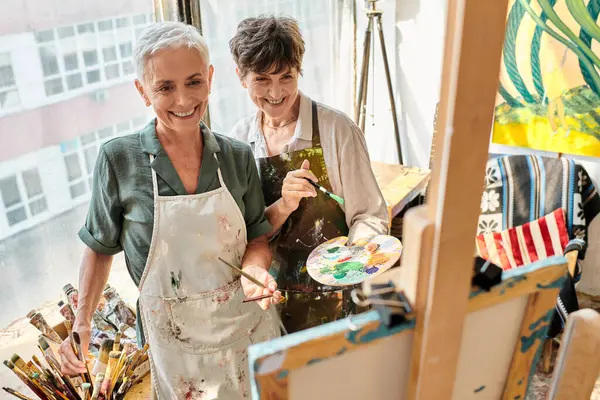 Cheerful mature women with paintbrushes looking at blurred easel during master class in art studio — Stock Photo