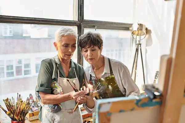 Middle aged female artists in aprons choosing paintbrushes during master class in art studio — Stock Photo