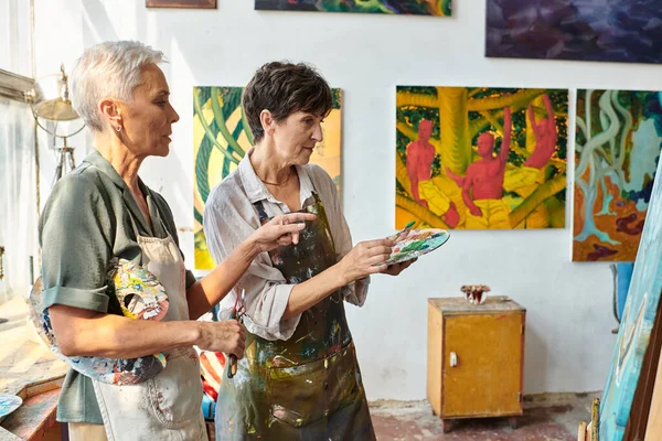 Skilled female artist pointing at easel near mature woman with palette during master class in studio — Stock Photo