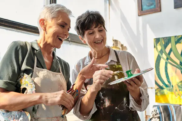 Cheerful female artist helping mature woman mixing paints on palette, master class in workshop — Stock Photo