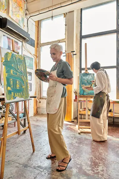 Creative mature woman in apron painting on easel near female friend in modern spacious art workshop — Stock Photo