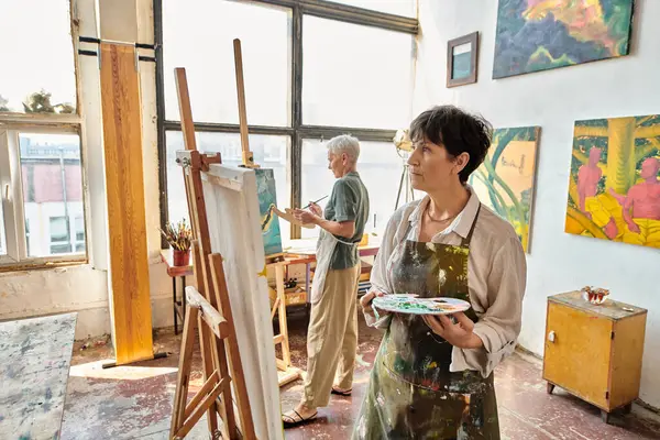Thoughtful mature woman in apron looking at easel near female friend in modern spacious art workshop — Stock Photo