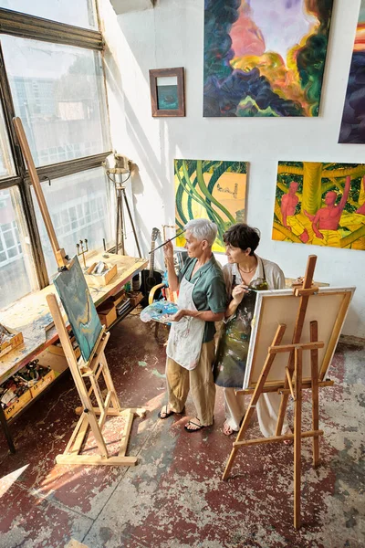 Високий кут зору зрілих друзів-жінки в фартухах живопис на мольбертах в сучасній просторій майстерні — стокове фото