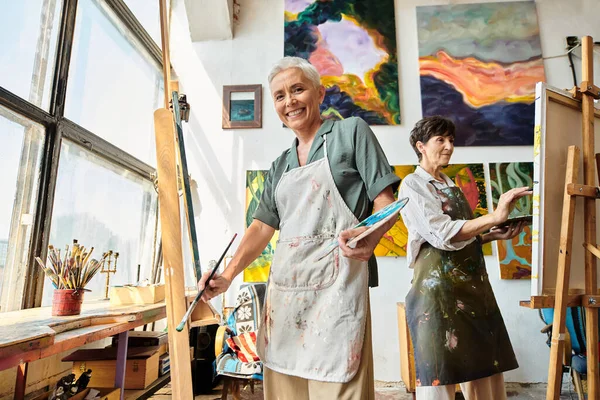 Überglückliche reife Frau in Schürze blickt in moderne geräumige Werkstatt in die Kamera, künstlerisches Hobby — Stockfoto