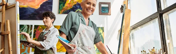 Happy mature woman smiling at camera near easel in workshop, artistic hobby, horizontal banner — Stock Photo