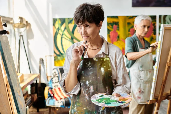 Thoughtful mature female artist with palette looking at easel in art workshop, creative process — Stock Photo